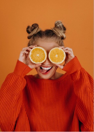 A photo of Sophie in an orange shirt, against an orange backdrop, holding orange slices in front of her eyes.