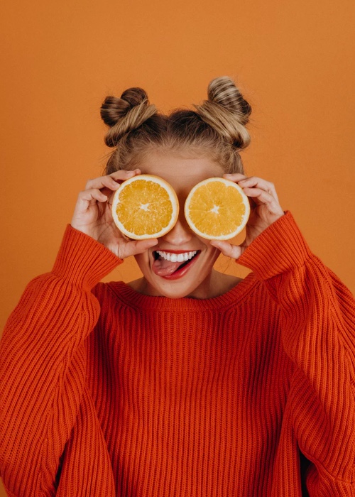 A portrait of Sophie in an orange sweater holding orange slices over her eyes